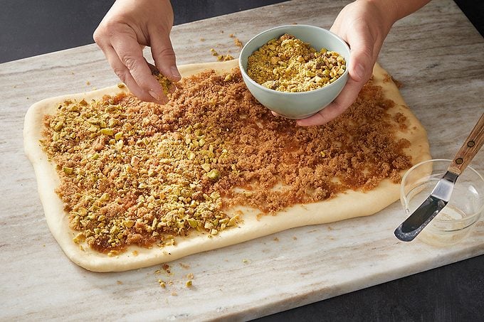 A person sprinkles chopped nuts and brown sugar over a rectangular sheet of dough on a marble surface, preparing it for baking.