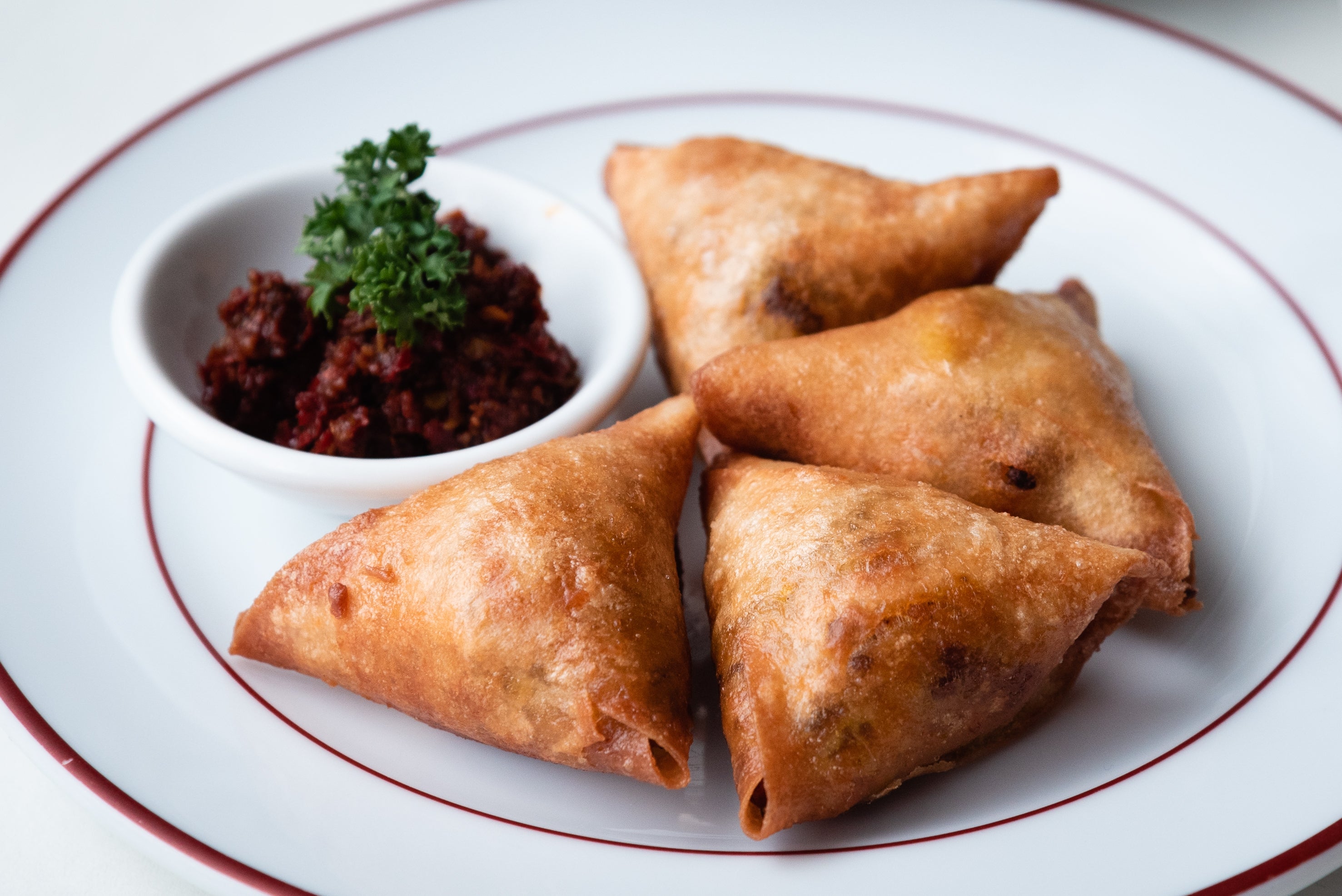 Four samosas with a side of chutney on a white plate, garnished with parsley