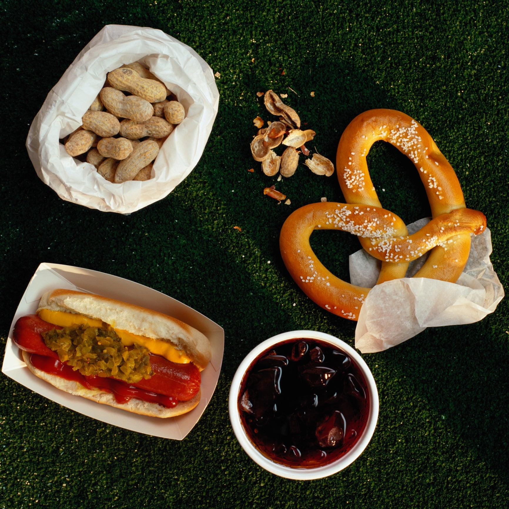 Hot dog with relish, soft pretzel, shelled peanuts, and a cup of soda on grass, resembling a classic sports stadium snack setup