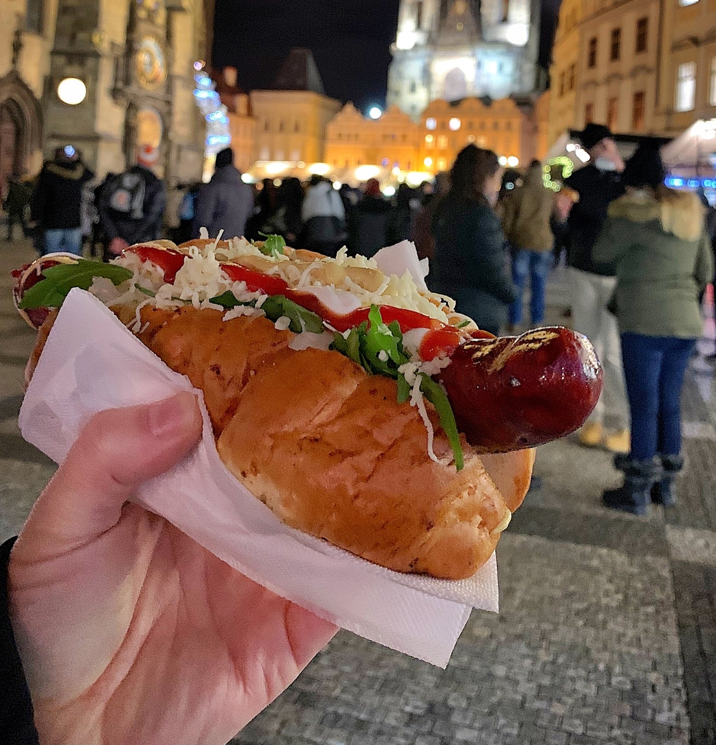 A hand holding a hot dog with toppings in a lively, historic town square at night, with a cathedral in the background