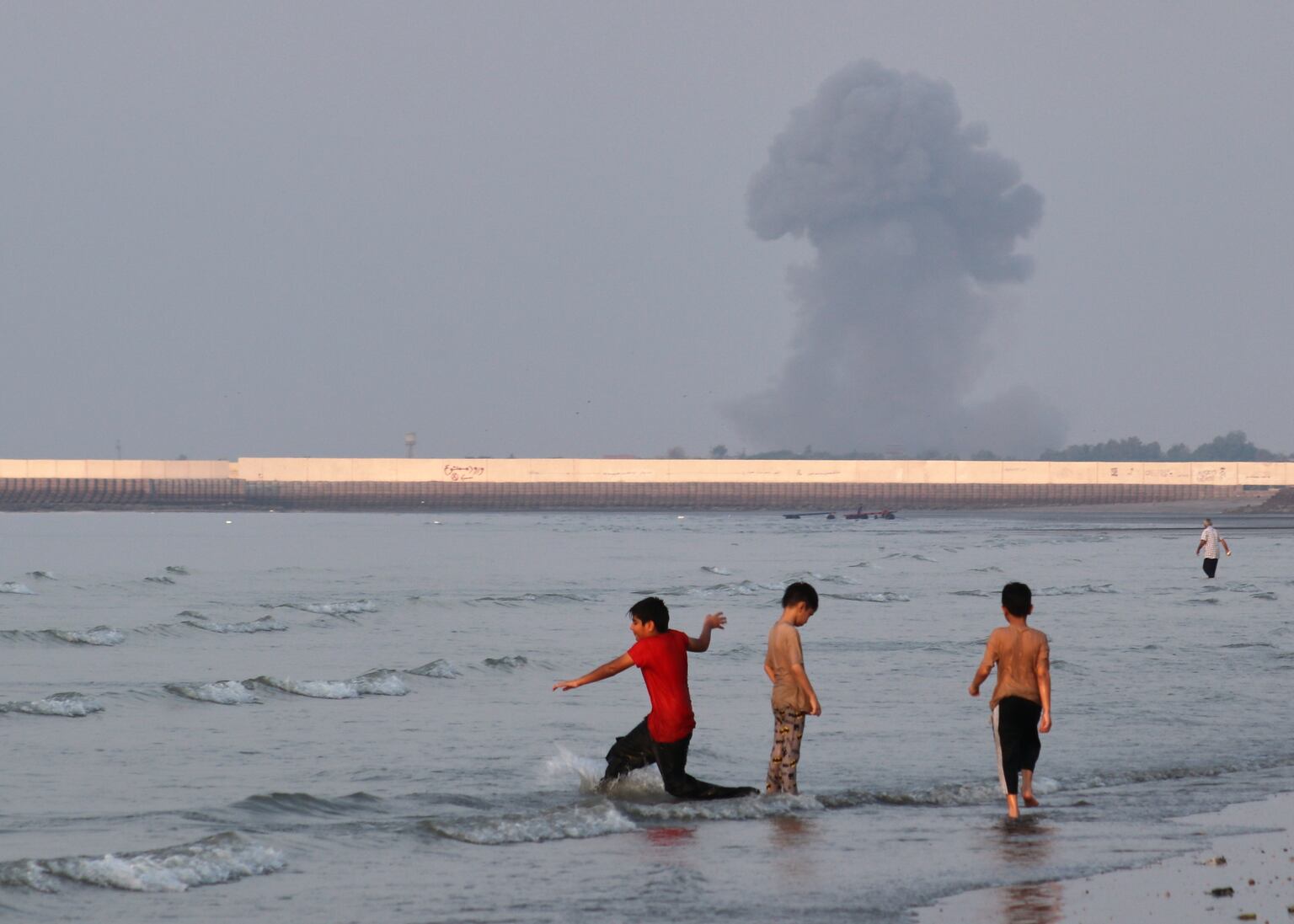 Three boys play in the shallow waters of the Strait of Hormuz, as a plume of smoke rises from an explosion in the background, off Bandar Abbas, Iran (Razieh Poudat/ISNA/AP)