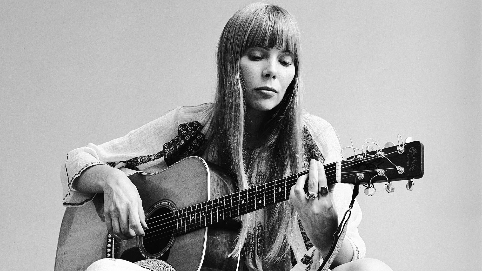 Joni Mitchell seated on the floor playing acoustic guitar, November 1968. This image is from a shoot for the fashion magazine Vogue. Mitchell wears a loose-fitting white dress.