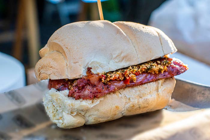 Close-up of a sandwich with sausage and a topping of diced vegetables or relish on a crusty roll. The sandwich is on a sheet of paper