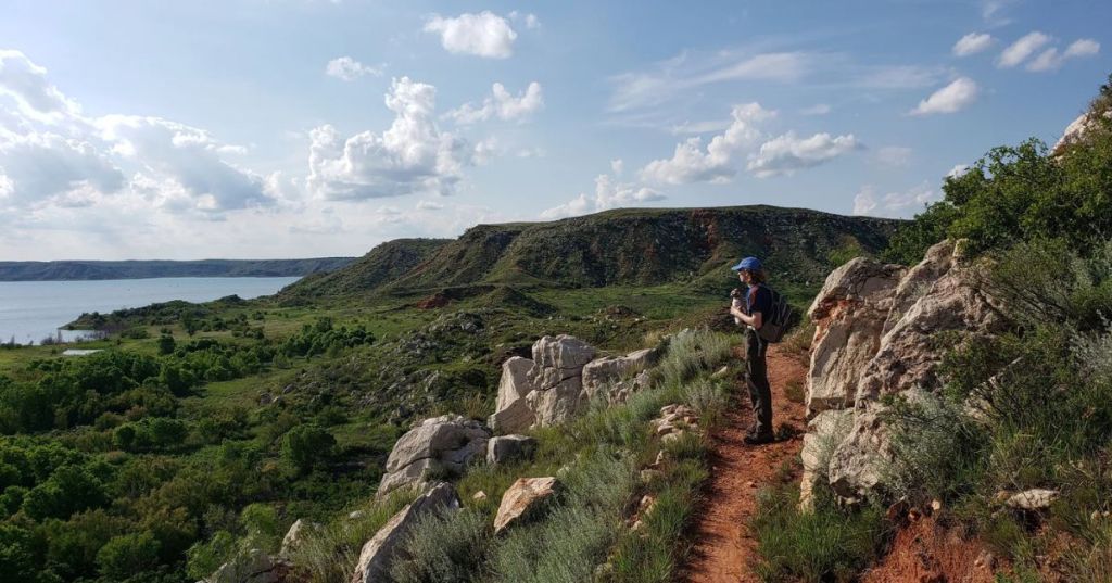 A hiker on a rocky trail overlooking Lake Meredith in Texas, featuring lush hills and calm waters in the distance.