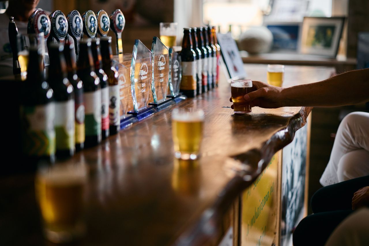 A bar counter with several beer bottles a hand holding a glass of beer and beer taps in the background