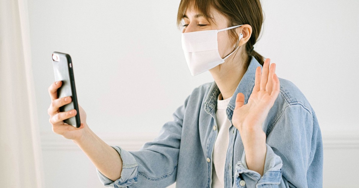 A woman wearing a face mask waves while video calling indoors, symbolizing remote connection during COVID-19.