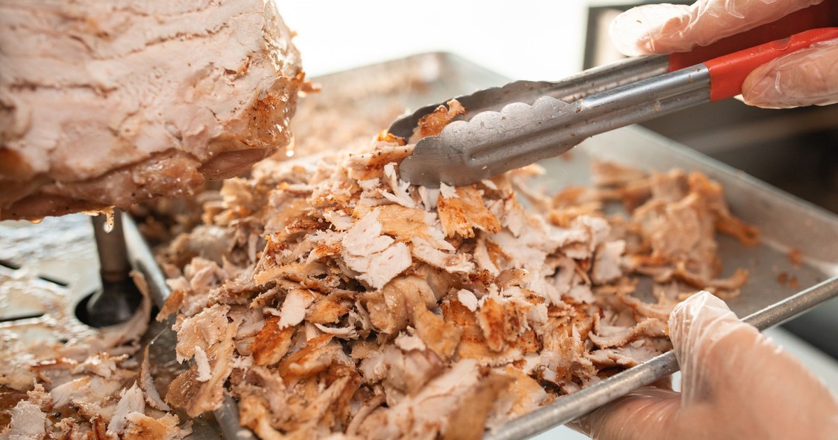Freshly sliced shawarma meat on a tray being prepared with tongs in a kitchen setting.