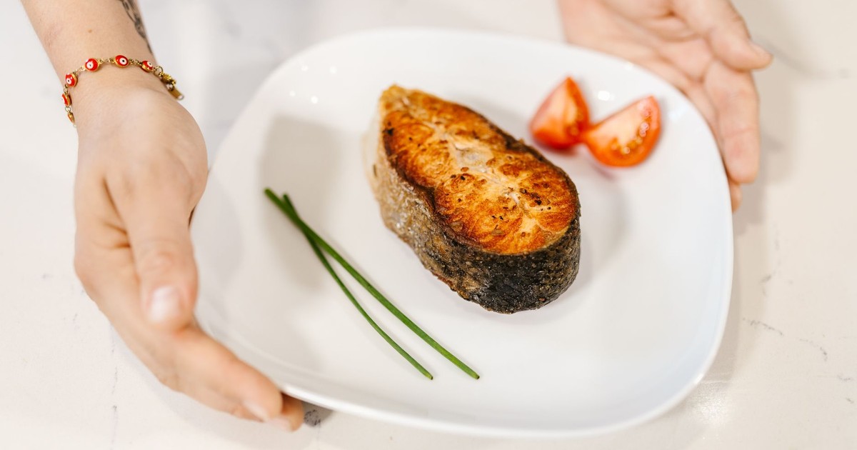 Close-up of grilled salmon served with tomatoes and chives on a plate, held by tattooed hands.