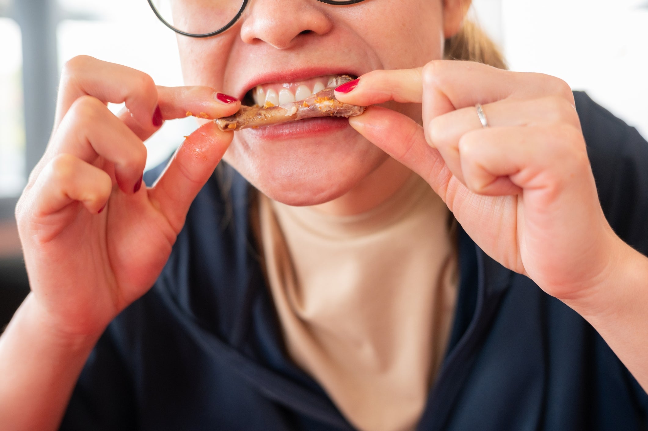 Person biting into a small fish, displaying teeth, with glasses, red nails, and a ring visible