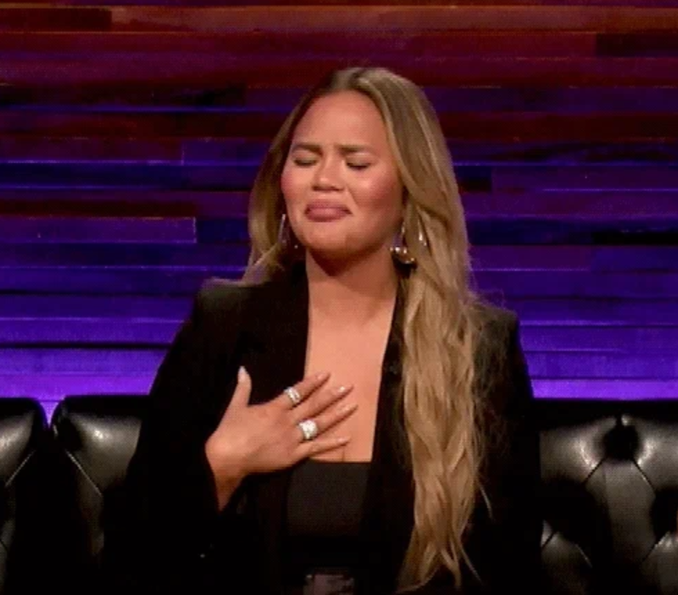 A person with long hair and large earrings is seated, making an expressive face with eyes closed and a hand on their chest
