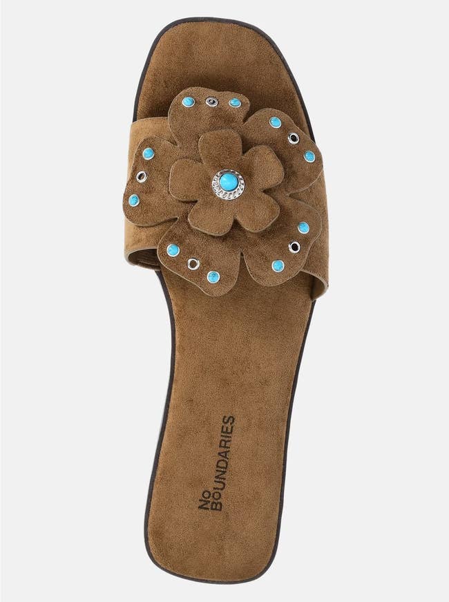 Brown slide sandal with large flower embellishment on strap, featuring blue and silver studs. Brand name