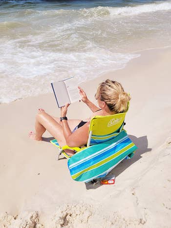 Person relaxing on a beach, sitting in a striped beach chair, reading a book near the shore