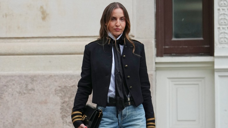 A woman wearing an unbuttoned military uniform jacket in front of a white wall.