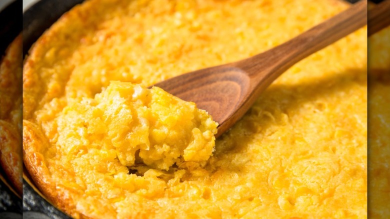 A wooden spoon scoops out of a casserole dish filled with baked corn pudding