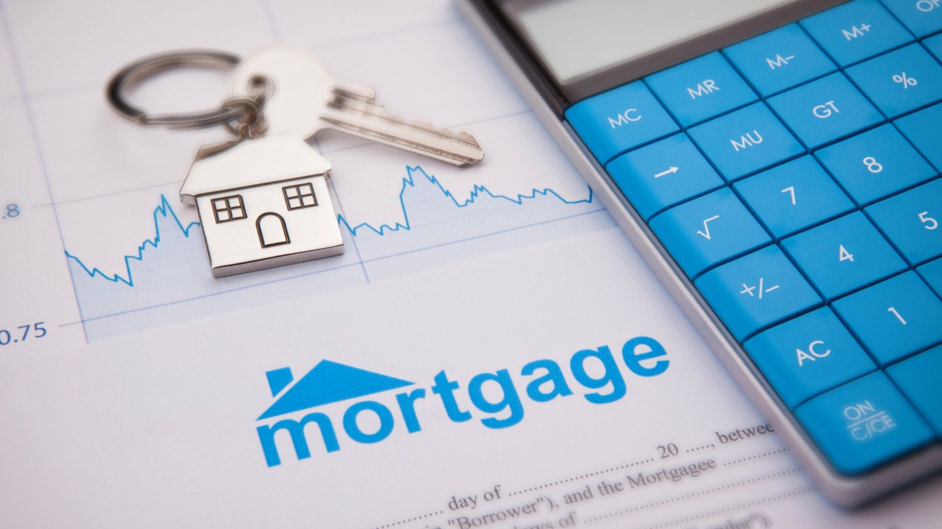 A close-up shot shows a silver house-shaped keychain with a key resting on a white document. The document features the word 'Mortgage' in prominent blue text with a small house icon, and a blue line graph with numeric axes (0.75, 0.8) indicates market fluctuations. A blue calculator is partially visible on the right side of the frame.