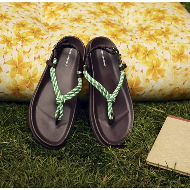Sandals with braided green straps on grass, next to a floral-patterned cushion and a closed book. Suitable for casual outdoor wear