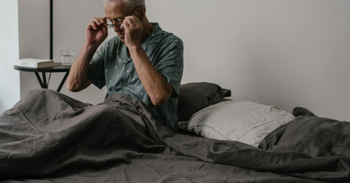 Senior man adjusts glasses while sitting in bed, expressing morning routine and daily life.
