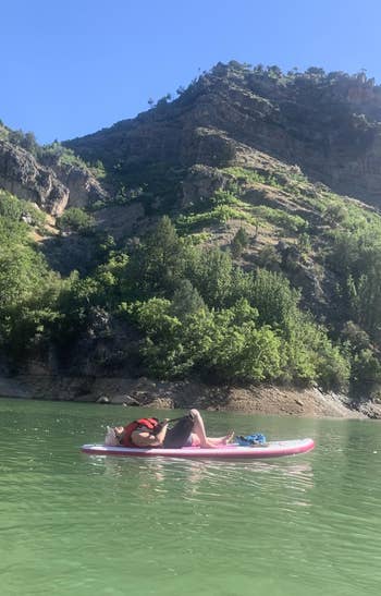 A person relaxes on a paddleboard in a calm lake surrounded by lush, green hills under a clear sky