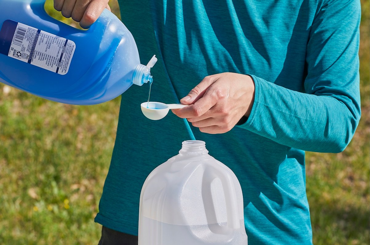 Woman measures dish soap into a tablespoon , preparing to add it to a gallon jug of water.