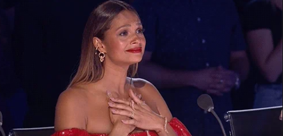 An individual in elegant off-the-shoulder attire sits, hands clasped in emotion, possibly on a talent show judging panel