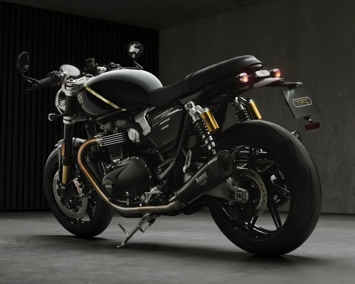Black Triumph Speed Twin 1200 TFC motorcycle with gold accents and Akrapovic exhaust on a gray floor.