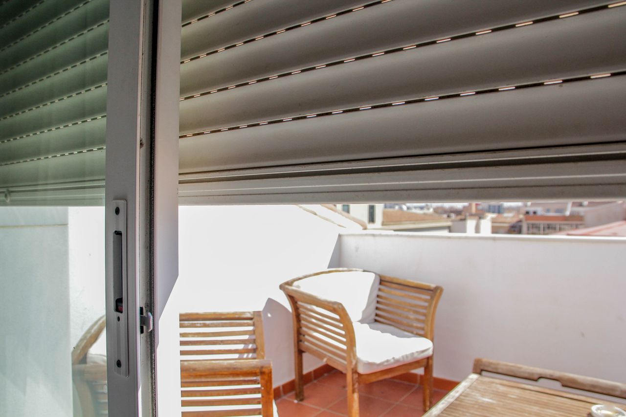 An exterior view of a balcony with wooden chairs and a partial open window shutter