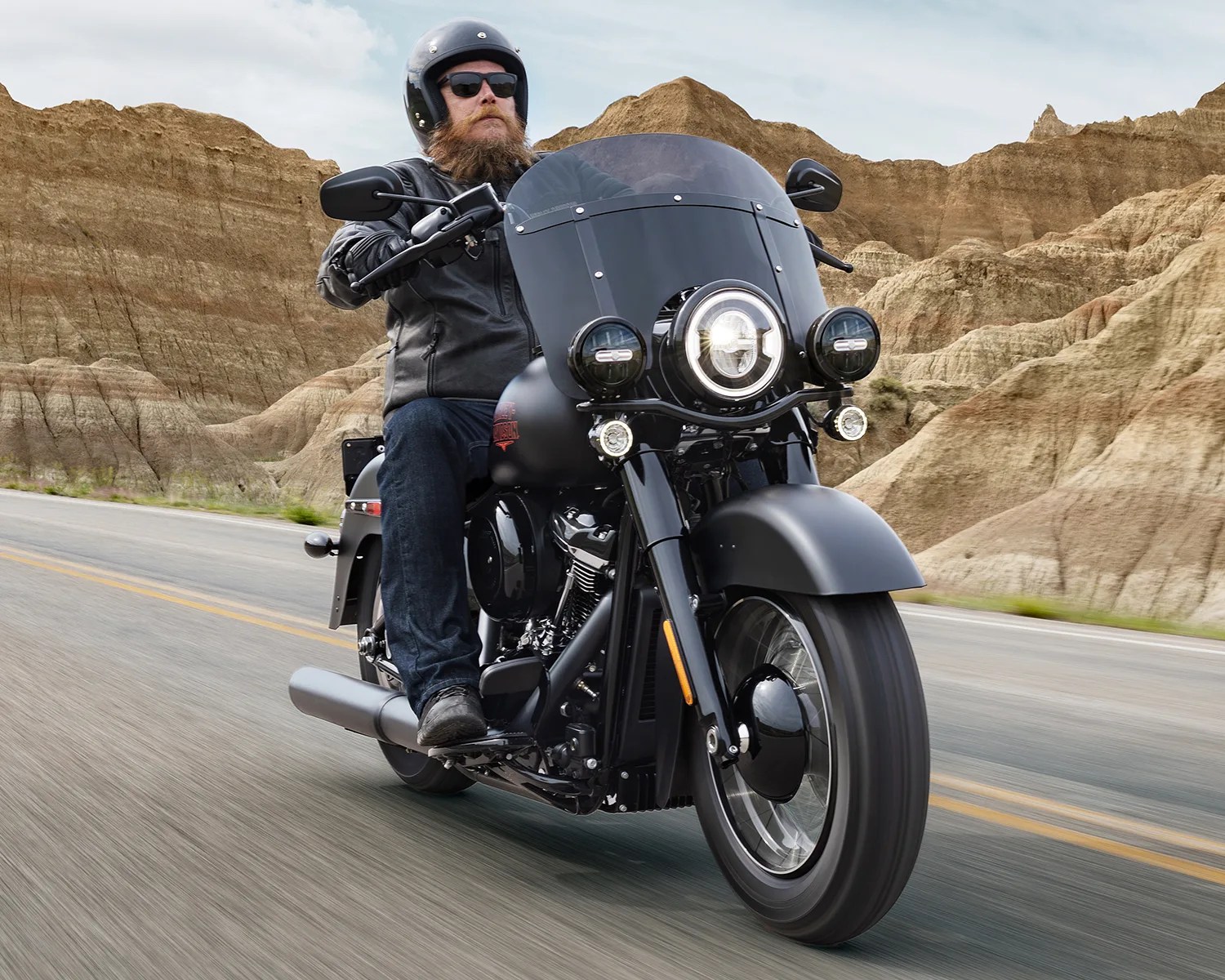 Man wearing black helmet and leather jacket riding a black motorcycle on a desert road with rocky hills in the background.