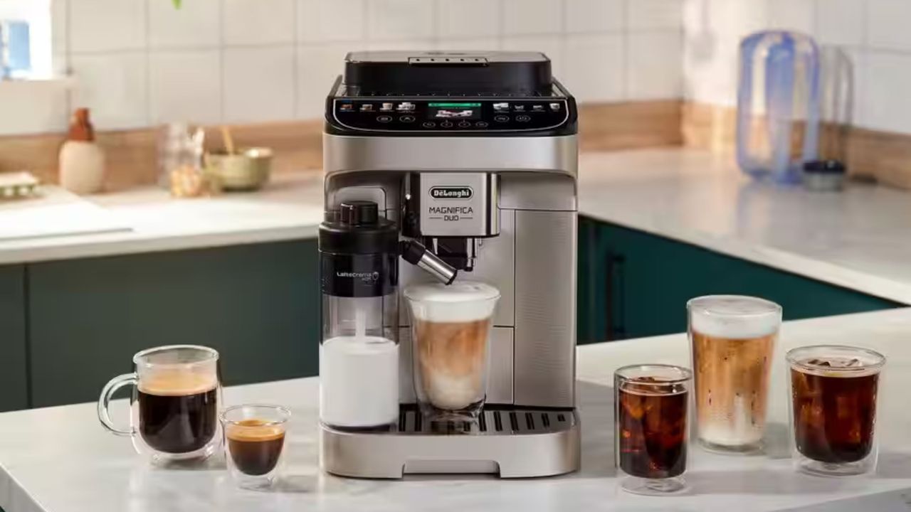 A De'Longhi coffee machine in black and stainless steel with a glass of iced coffee on the drip tray beside an automatic milk dispenser.