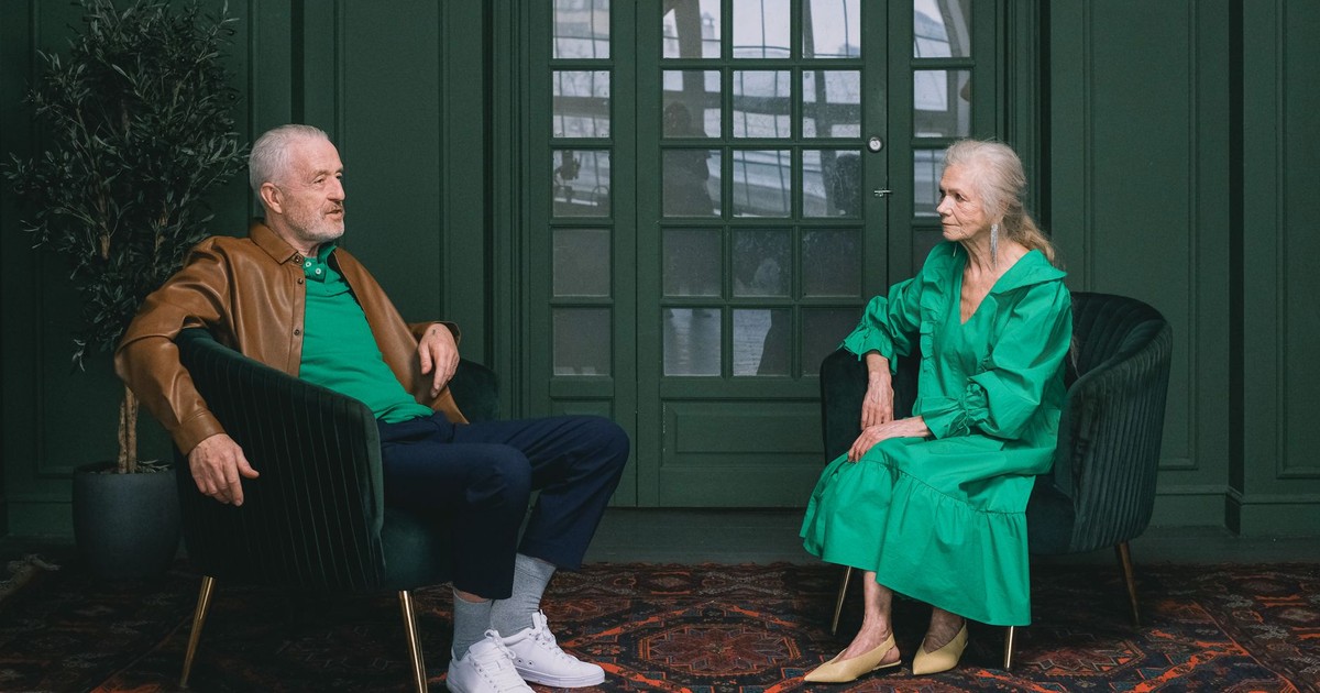 Elderly man and woman in green-themed room engaged in conversation, seated on armchairs.
