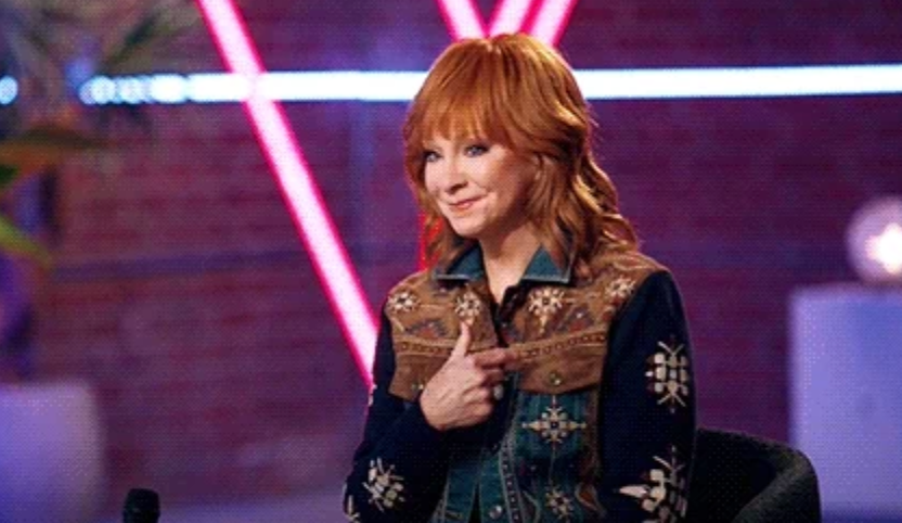 Person seated with shoulder-length hair, wearing a patterned jacket with floral details, gesturing towards themselves. Background has soft lighting effects