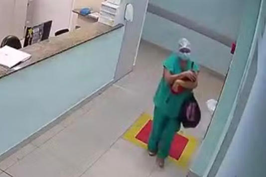 A woman wearing scrubs holding a baby and a black bag in a hospital
