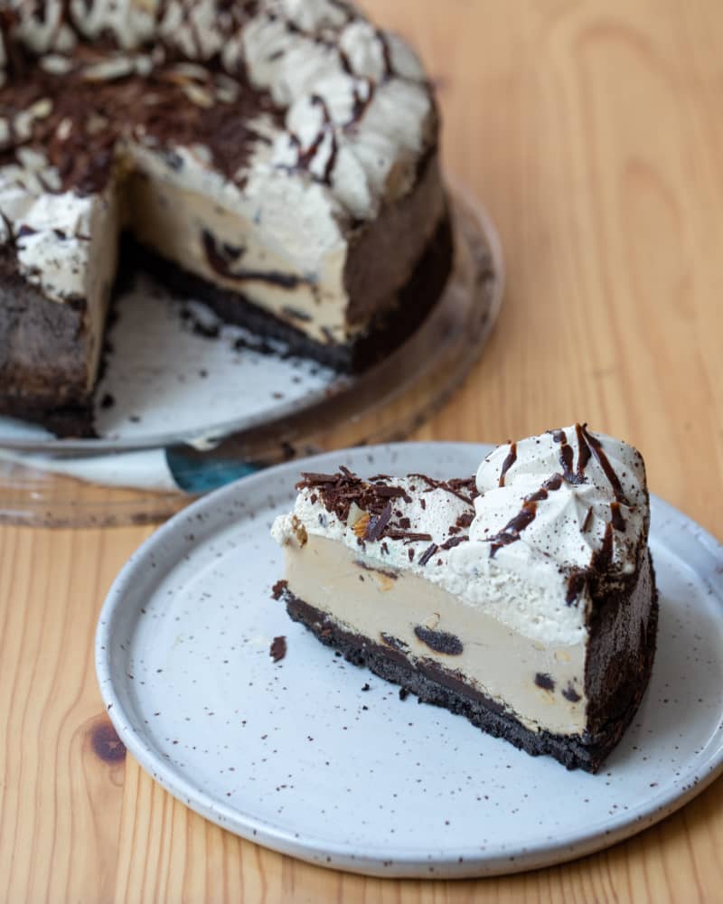 Slice of ice cream cake with chocolate crust, whipped cream, and chocolate shavings on a speckled plate.