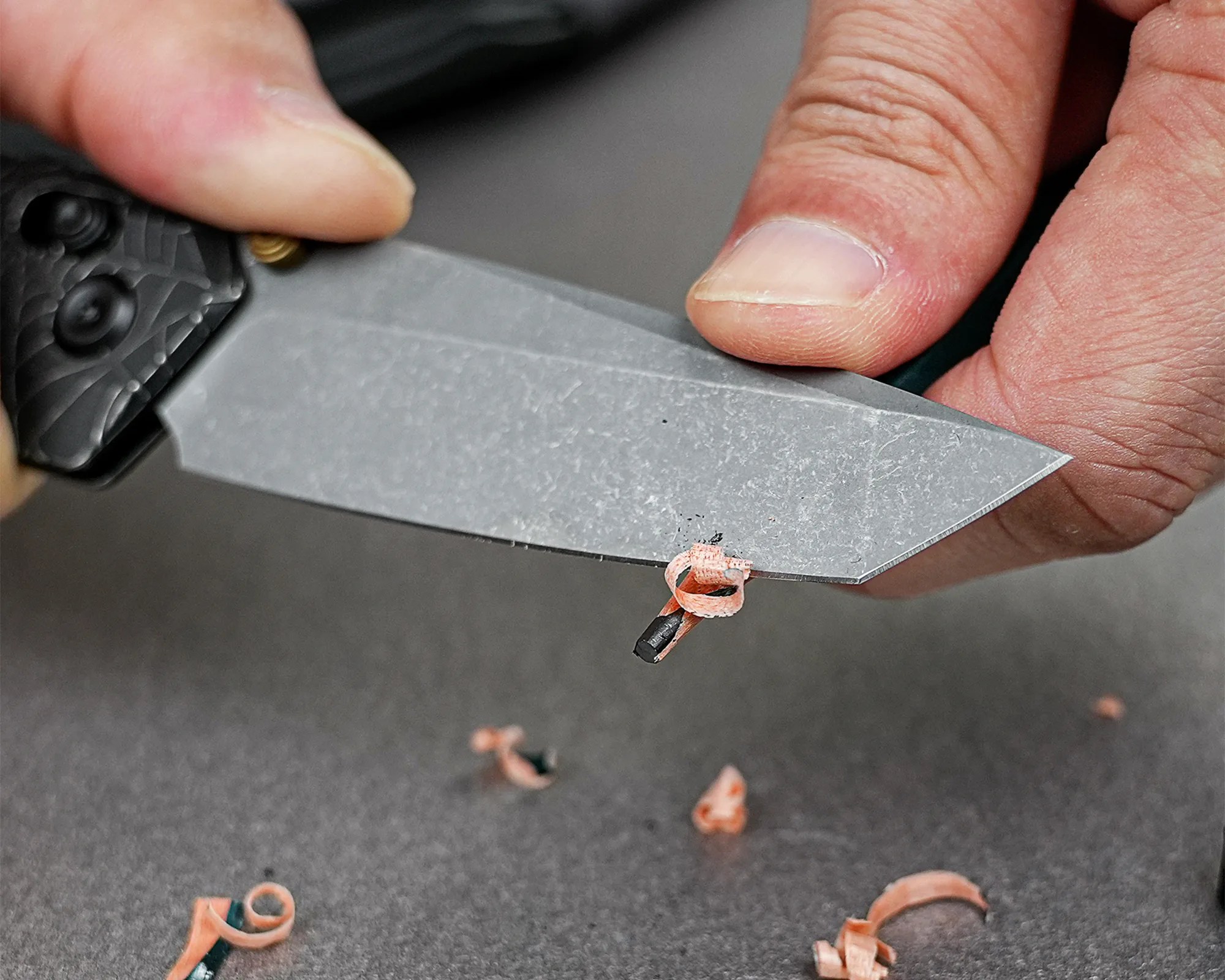 Hand holding a black folding knife with a stonewashed blade shaving a small wood curl.