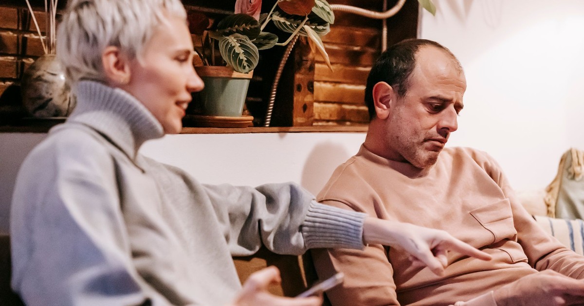 Side view of positive adult woman in stylish sweater sitting on couch using smartphone and pointing at screen of tablet in hands of focused ethnic husband