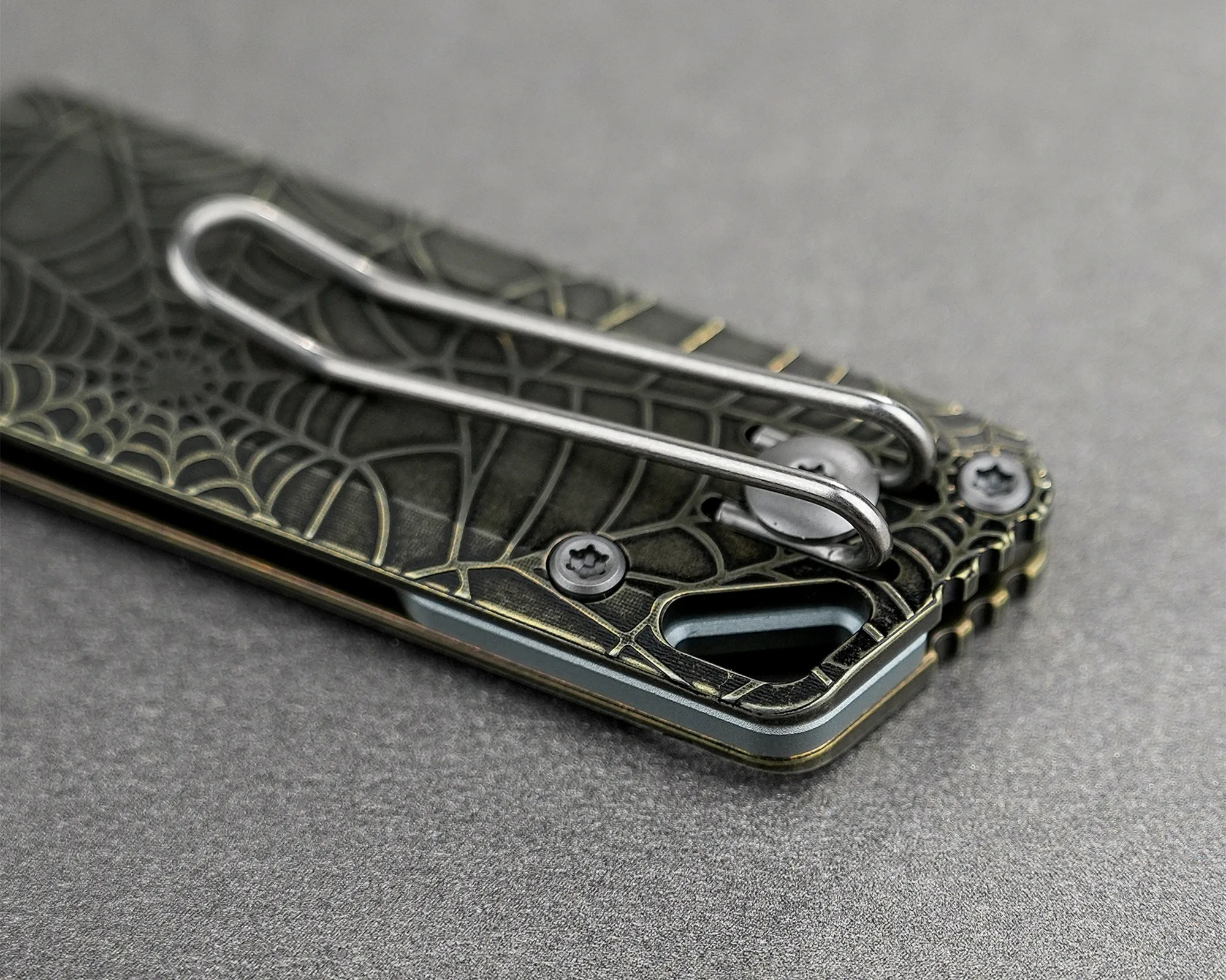 Close-up of a black and gold spiderweb-patterned metal object with a silver wire clip and screws on a gray surface.