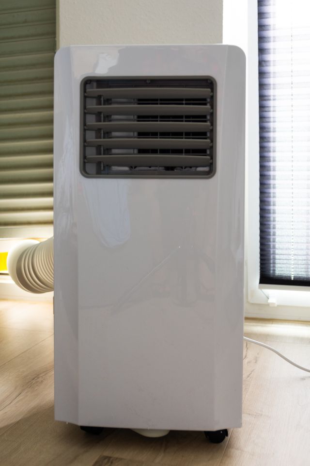 A portable air conditioner on the floor with a vent and exhaust hose located near a window with blinds