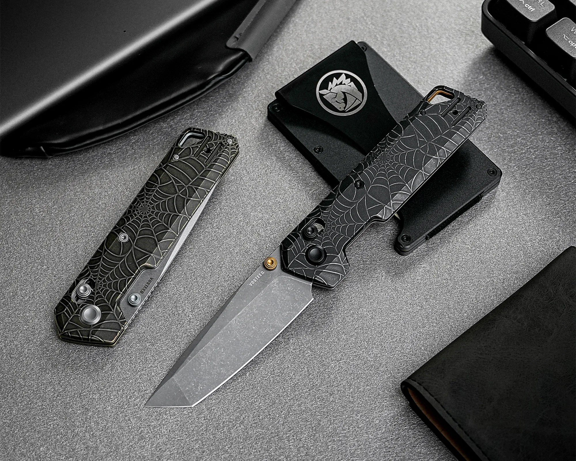 Two black folding knives with spiderweb-patterned handles on a gray textured surface next to a black wallet and a black cardholder with a silver logo.