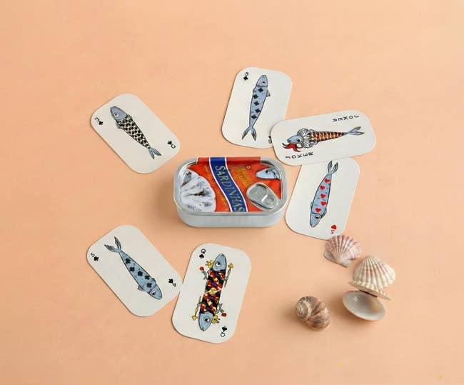 A sardine tin surrounded by playing cards with fish designs and seashells, suggesting a whimsical, nautical-themed shopping item display