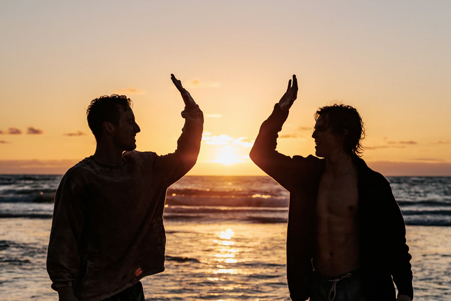 Two men high-fiving on a beach at sunset, silhouetted against the orange sky.