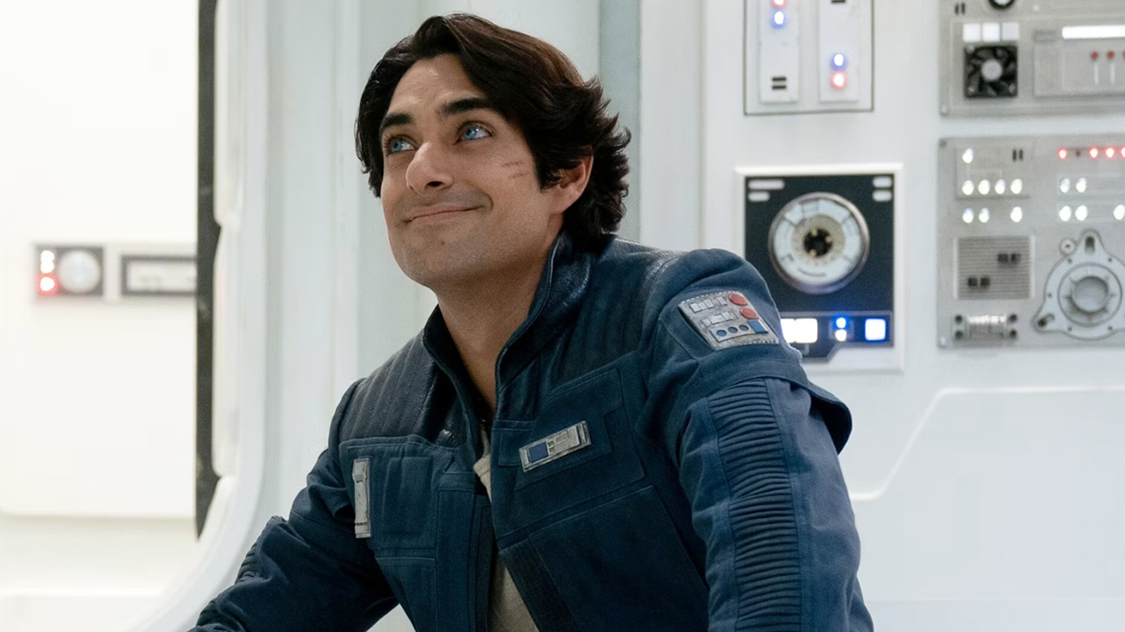 A man with bright blue eyes and dark hair is sitting in a futuristic room. He is looking off into the distance with a smile on his face.