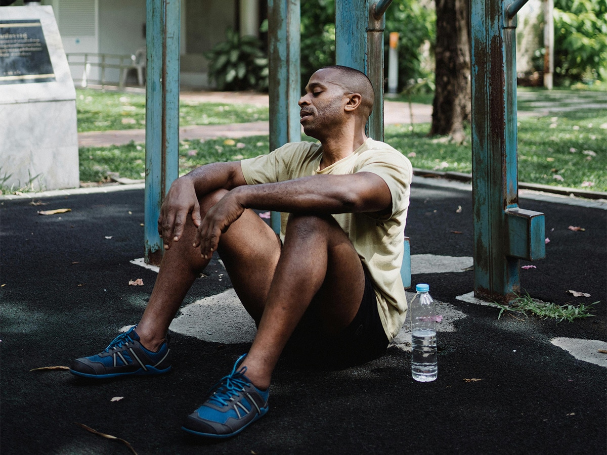 Man resting on gym floor outdoors, wearing a yellow shirt and blue sneakers, with a water bottle nearby.