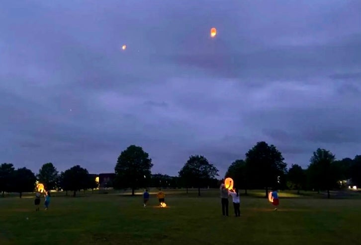 Eddie's friends releasing lanterns into the night sky in a grassy open area with trees silhouetted in the background