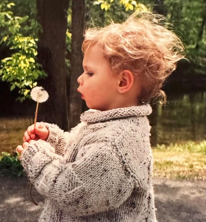 Eddie as a toddler blowing a dandelion outdoors while wearing a cozy knitted sweater