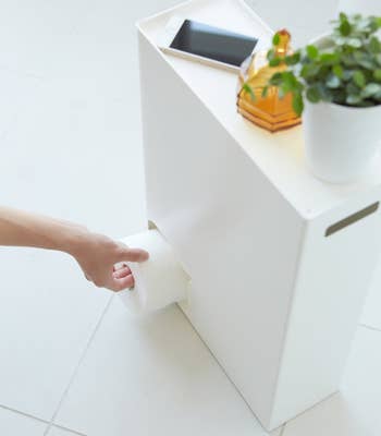 Hand pulling toilet paper from a sleek, modern storage unit with a phone and plant on top