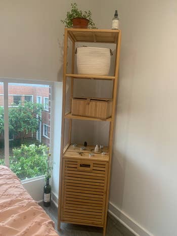 Tall wooden shelf unit with plants, decorative items, and a wicker basket, positioned by a window with a view of trees outside