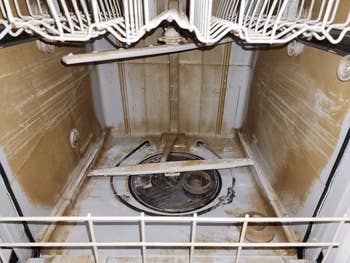 Inside view of a worn dishwasher showing rust stains and mineral buildup on racks and spray arms, indicating the need for deep cleaning or replacement