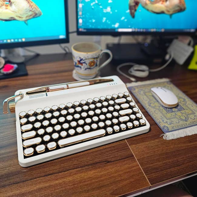 A vintage-style mechanical keyboard on a desk with a mouse, ornate mug, and two monitors in the background