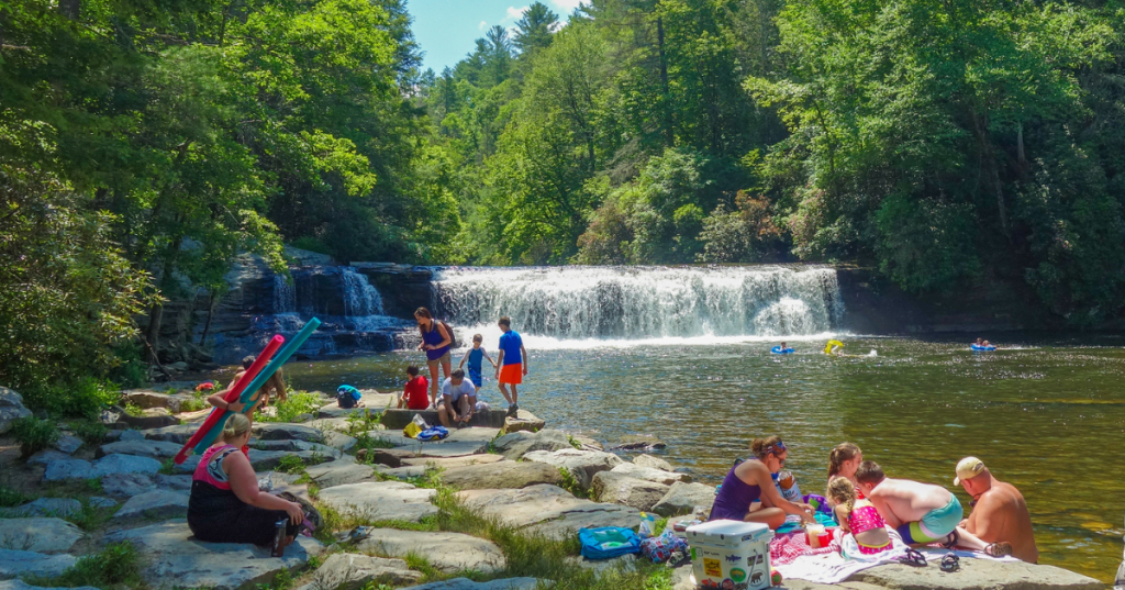 Visitors wading and swimming in the shallow waters at the base of Hooker Falls on a sunny summer day.