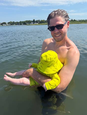 A parent in sunglasses stands in the water holding a baby dressed in a sun hat and swimwear