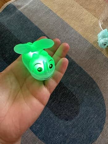 A smiling animal-shaped toy with propeller eyes, glows, held in a hand over a textured surface